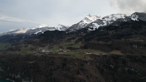 Aerial-pan-across-homes-dotting-grassy-hillside-on-plateau-above-Lake-Walen-with-snow-covered-mountain-peaks