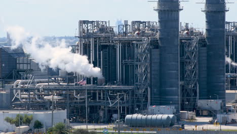 Explore-the-immense-scale-of-clean-energy-natural-gas-plant-with-billowing-white-smoke,-framed-by-the-USA-Flag-waving-in-the-video-background,-reflecting-energy-innovation-and-national-ethos