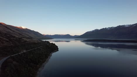 Vuelo-Aéreo-Sobre-El-Lago-Wakatipu-Con-La-Carretera-Al-Lado-Y-Rodeado-De-Montañas-Cubiertas-De-Nieve-Que-Se-Reflejan-En-El-Lago-Justo-Después-Del-Atardecer-En-Nueva-Zelanda