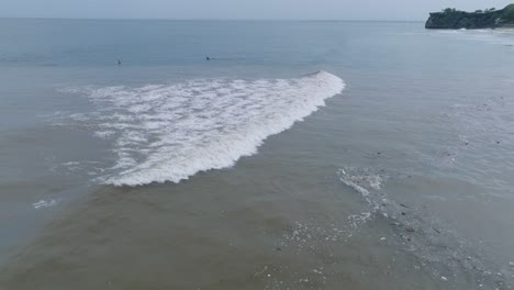 Low-altitude-pan-down-drone-shot-of-polluted-ocean-filled-with-floating-trash-and-debris-and-muddy-sewage-river-runoff-with-surfers-in-the-background-in-Bali-Indonesia