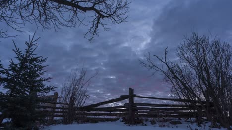 Timelapse-of-sunrise-by-trees-and-fence-in-snowy-rural-Canada,-tilt-down