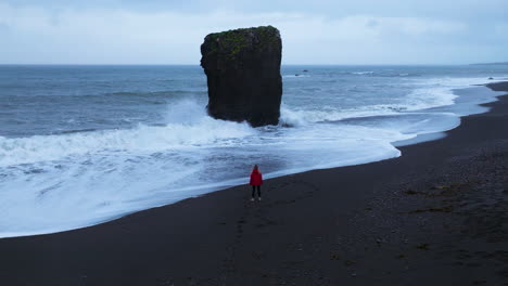 Woman-In-Red-Jacket-Standing-On-Laekjavik-Black-Sand-Beach-In-Oceanside,-Iceland