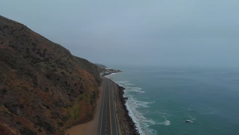 Slow-pull-back-establishing-drone-shot-as-car-pulls-out-onto-Pacific-Highway-One,-near-Malibu-on-a-grey-and-foggy-morning