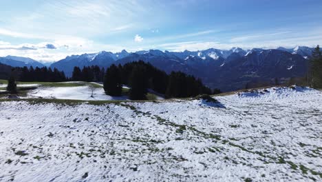 Drohnenflug-über-Einem-Schneefeld,-Das-Grüne-Grashügel-Und-Einen-Bauernhof-Auf-Den-Bergen-Enthüllt,-Mit-Herrlichem-Blick-Auf-Schneebedeckte-Berge-An-Einem-Sonnigen-Tag-Mit-Blauem-Himmel