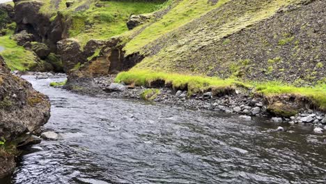Steadily-running-river-winding-along-green-mountainside-slopes,-wide-static-shot