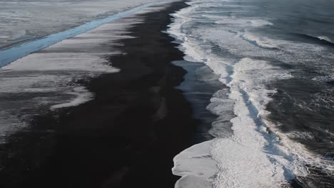 Slow-drone-flyover-black-beach-with-foamy-waves-at-sunset