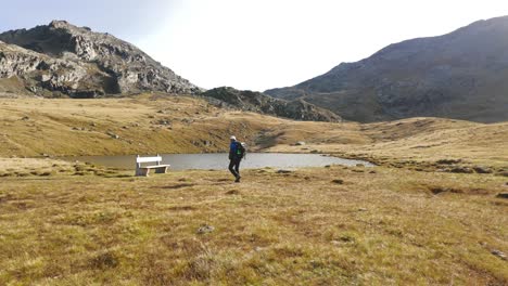 Male-Hiker-With-Backpack-Walking-Across-Grassland-Towards-Bench-Beside-Lake-At-Valmalenco,-Italy-On-Cleat-Sunny-Day