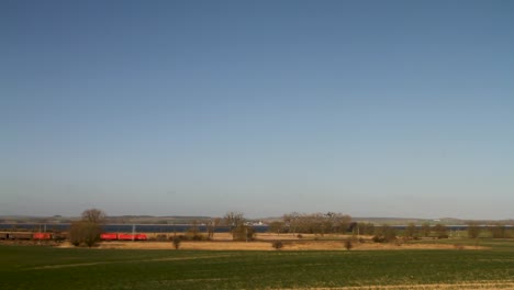 Red-train-crossing-rural-landscape-with-clear-blue-skies,-wide-shot-during-daytime