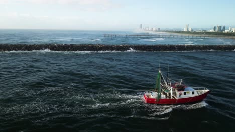 Ein-Fischtrawler-Läuft-In-Einen-Küstenhafen-Ein,-Im-Hintergrund-Sieht-Man-Die-Skyline-Einer-Stadt-Und-Surfstrände