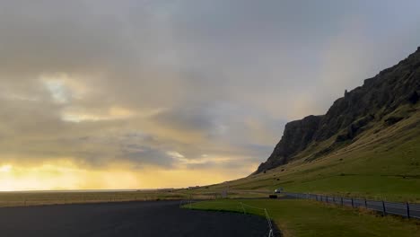 Wide-golden-hour-view-of-cars-on-tranquil-Icelandic-road,-majestic-cliffs-under-sunset-sky