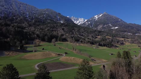 Vista-Aérea-De-La-Carretera-Y-Pequeñas-Cabañas-Con-Montañas-Cubiertas-De-Nieve-En-El-Fondo-En-Un-Hermoso-Día-Soleado-Con-Un-Cielo-Azul-Claro-En-Austria