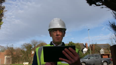 A-happy-senior-architect-examining-plans-of-a-large-building-on-a-tablet-on-a-construction-site-in-a-residential-street-with-traffic-on-the-road-in-the-background