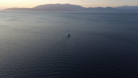 Einzelnes-Boot,-Das-Mit-Wellen-Auf-Dem-Meer-Schwimmt,-Geneigte-Luftaufnahme-In-Richtung-Sonnenbeschienener-Horizont-Und-Bergiger-Küste