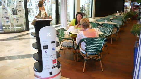 A-working-futuristic-AI-robot-waiter-glides-through-a-bustling-cafe,-its-digital-eyes-and-bowtie-adding-a-quirky-touch-to-the-dining-experience-utilizing-artificial-intelligence-and-automation