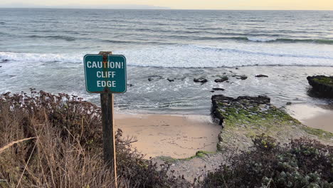 Caution-sign-by-the-sea-at-cliff's-edge-with-waves-rolling-in-at-dusk,-Santa-Cruz-overlooking-Monterey-Bay