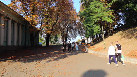 People-walking-in-a-park-towards-a-monument-The-Arch-of-Freedom-of-the-Ukrainian-People-in-Kyiv-city-center-Ukraine,-Arka-Svobody-Ukrayinsʹkoho-Narodu,-sunny-weather-and-autumn-trees,-4K-shot