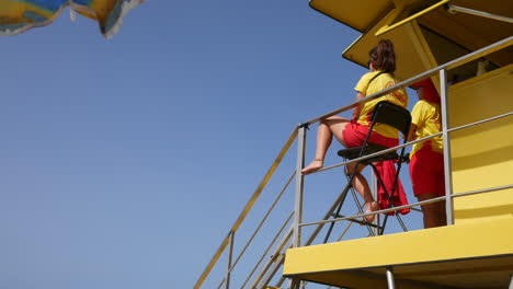 Socorristas-Vestidos-De-Rojo-Y-Amarillo-Inspeccionan-La-Playa-Desde-Lo-Alto-De-Una-Torre-Amarilla-Contra-Un-Cielo-Azul-Claro-Y-Profundo,-Vigilantes-De-La-Seguridad-Junto-Al-Mar