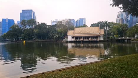 Lago-Del-Parque-De-La-Ciudad-Con-Fuente-En-Primer-Plano,-Rascacielos-Al-Fondo-Al-Atardecer,-Ambiente-Sereno,-Parque-Lumpini-En-Bangkok,-Tailandia