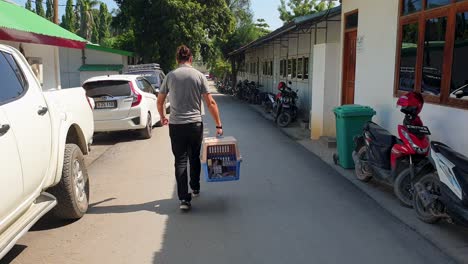 A-man-walking-down-street-carrying-cat-in-pet-carrier-box-after-vet-health-check-up-and-inspection-in-preparation-for-flying-and-travel-to-overseas-destination