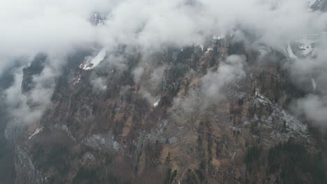 Panoramic-overview-of-wispy-tendrils-of-clouds-up-against-sheer-mountain-cliffs-covered-in-snow