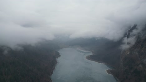 Thick-misty-clouds-settle-in-valley-shrouding-frozen-lake-or-reservoir-in-the-alps