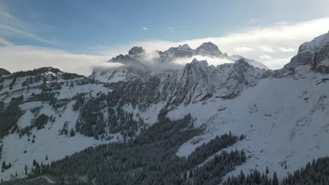 Panorama-Luftaufnahme-Von-Zerklüfteten-Felsrücken-Und-Schneebedeckten-Kiefernwäldern-Mit-Wolken,-Die-Sich-Um-Den-Berg-Sammeln