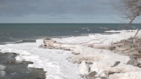 Icy-shoreline-at-Southampton-with-driftwood,-winter-waves-crashing,-and-a-cloudy-sky,-wide-shot