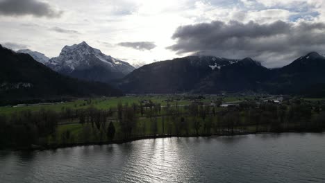 Sunlight-breaks-between-clouds-with-ray-of-light-shimmering-on-lake-and-farmland-fields,-epic-swiss-mountain-peaks