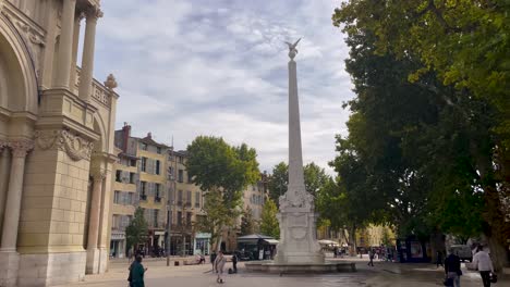 Ein-Sonniger-Tag-Auf-Einem-Belebten-Platz-In-Aix-en-Provence-Mit-Fußgängern-Und-Einem-Hoch-Aufragenden-Historischen-Denkmal