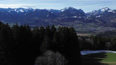 Drohnenflug-über-Wald-In-Wunderschöner-Berglandschaft,-Kleine-Hütte-Auf-Den-Hügeln-Mit-Schneebedeckten-Bergen-Und-Stadtbild-Im-Hintergrund