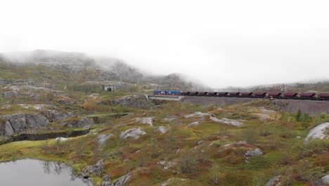 Aerial:-ore-train-in-Søsterbekk-stasjon,-close-to-the-border-between-Sweden-and-Norway-in-north-Lapland-crossing-a-bridge-on-a-lake-and-entering-a-tunnel-in-a-misty-landscape