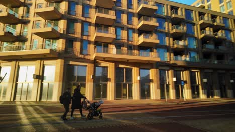 Couple-with-baby-passing-by-at-modern-urban-square-in-Amsterdam-Noord-with-low-morning-sunlight