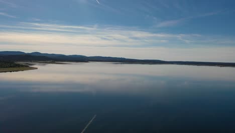 Wenn-Wir-Mit-Einer-Drohne-über-Einen-Stausee-Fliegen,-Sehen-Wir-Die-Spiegelung-Des-Blau-weißen-Himmels-Mit-Wolken-Im-Wasser,-Als-Wäre-Es-Ein-Spiegel.-Da-Ist-Das-Kielwasser-Eines-Flugzeugs-Und-Wir-Sehen-Ein-Ufer,-Alles-Ist-Ruhig