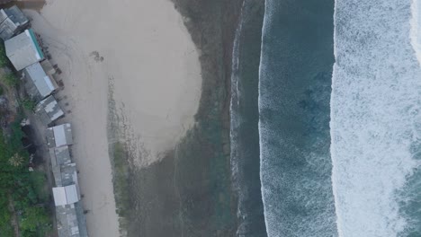 Top-down-drone-shot-over-tropical-Balangan-Beach-peninsula-in-Uluwatu-Bali-Indonesia-with-turquoise-waves-crashing-over-coral-reef-and-rustic-shacks-lining-beachfront