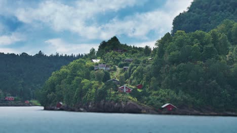 Miniaturized-tidy-red-wooden-cabins-and-boathouses-scattered-along-the-forest-covered-fjord-coast