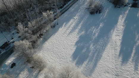Snowy-landscape-with-a-car-traveling-along-a-road,-trees-lining-the-path,-casting-long-shadows-in-the-morning-light,-aerial-tracking-shot