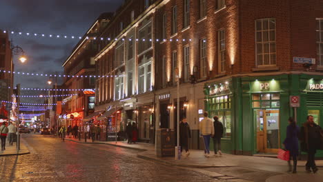 Pintoresca-Ciudad-Con-Gente-Caminando-Por-Calles-Adoquinadas-Por-La-Noche-En-Dublín,-Irlanda