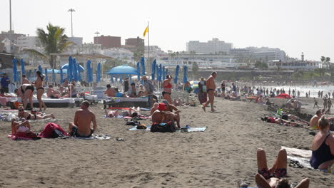 Crowded-sandy-beach-scene-with-a-yellow-flag,-sunbathers,-blue-umbrellas,-a-gentle-surf,-and-vacation-vibes-under-a-hazy-blue-sky-at-costa-adeje-tenerife