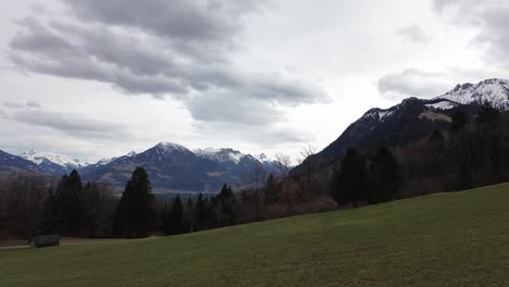 Luftaufnahme-Einer-Drohne,-Die-An-Einem-Bewölkten-Tag-In-Vorarlberg,-Österreich,-Durch-Ein-Grasfeld-Mit-Schneebedeckten-Bergen-Im-Hintergrund-Fliegt