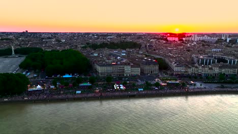 People-gathering-on-the-Garonne-river-shore-with-Ferris-Wheel-in-Place-des-Quinconces-Bordeaux-France-during-sunset,-Aerial-pan-left-reveal-shot