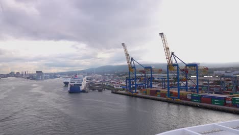 Cruise-Ship-Sailing-by-Cargo-Terminal-of-Belfast-Harbour,-Cranes-and-Shipping-Containers