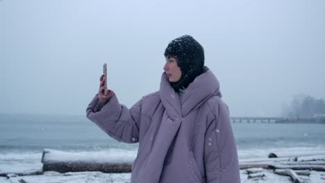 Woman-in-Warm-Clothing-Takes-Picture-with-Smartphone-on-Snowy-Beach
