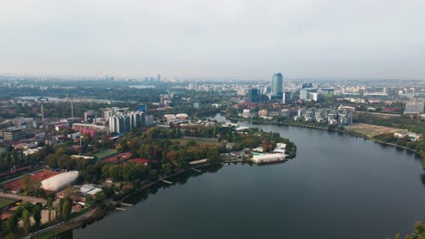 Lago-Floreasca-Con-Horizonte-De-Bucarest,-Aguas-Serenas,-Paisaje-Urbano,-Día-Nublado,-Paisaje-Urbano-Como-Telón-De-Fondo,-Vista-Aérea