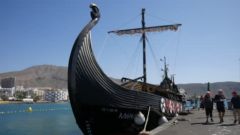 Viking-ship-tourist-replica-docked-in-a-modern-harbor,-tourists-walking-by,-with-cityscape-and-mountains-in-background