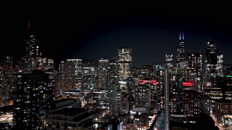 Aerial-view-backwards-over-the-night-lit-cityscape-of-River-north,-Chicago,-USA