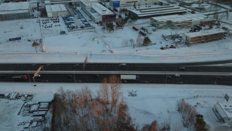 An-overhead-view-of-a-major-road-in-the-snow-clad-district-of-Dąbrowa-in-Gdynia,-showing-vehicles-moving-amidst-a-wintry-setting,-bordered-by-industrial-structures-and-bare-tree