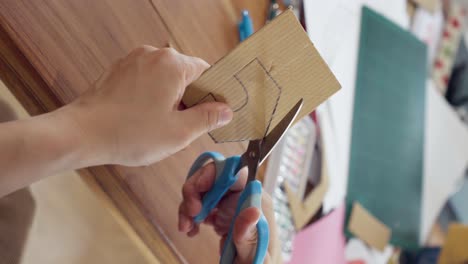 Close-up-of-hand-using-scissors-cut-a-house-on-cardboard
