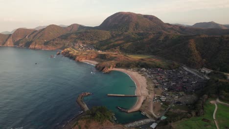 Aerial-drone-landscape-of-japanese-blue-sea-bay-in-kyoto-kyotango-island-summer-coastline,-white-sand-beach-with-nearby-village,-Japan-during-daylight,-mountain-range-background