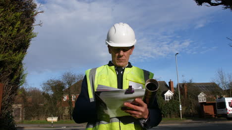 Low-Angle-Portrait-Eines-Leitenden-Bauleiters-Eines-Architekten-In-Einer-Wohnstraße-Mit-Verkehr-Und-Häusern,-Der-Sich-Die-Unterlagen-Zur-Gebäudeinspektion-Ansieht