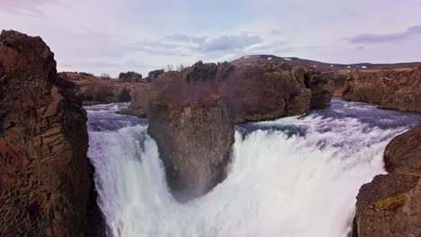 Flyover-twin-waterfalls-flowing-around-rocky-terrain-in-Iceland